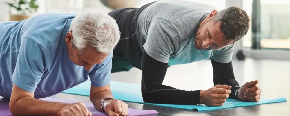Älterer Mann und Physiotherapeut führen gemeinsam eine Plank-Übung in einem hellen Trainingsraum durch – Physiotherapie Leipzig.
